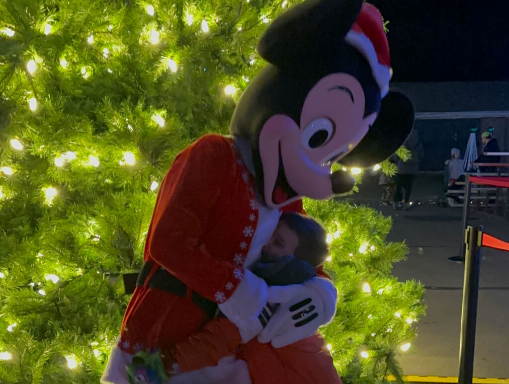 a young boy in an orange and gray coat hugs someone dressed as Mickey Mouse wearing a Santa hat and coat