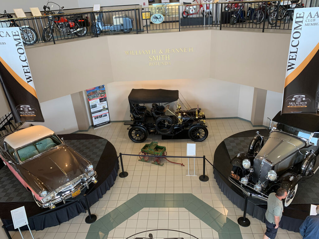 Rotunda of the AACA Museum featuring three antique cars on the first floor and a row of motorcycles and mopeds on the second floor