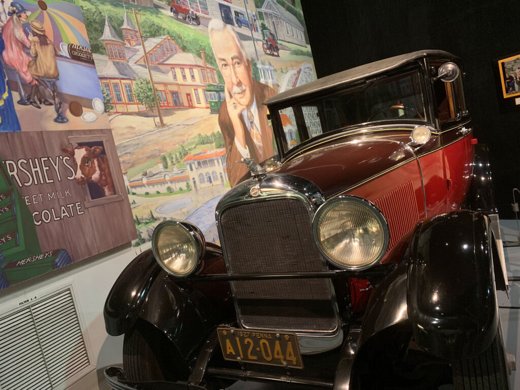 1920s era car in two-tone dark pink in front of a mural of Milton Hershey at the AACA Museum