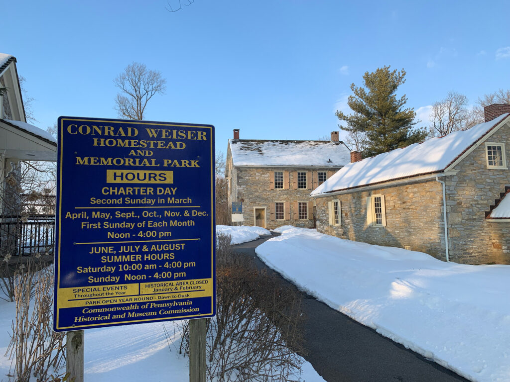 Blue sign with hours for the Conrad Weiser Homestead in front of  historic stone buildings