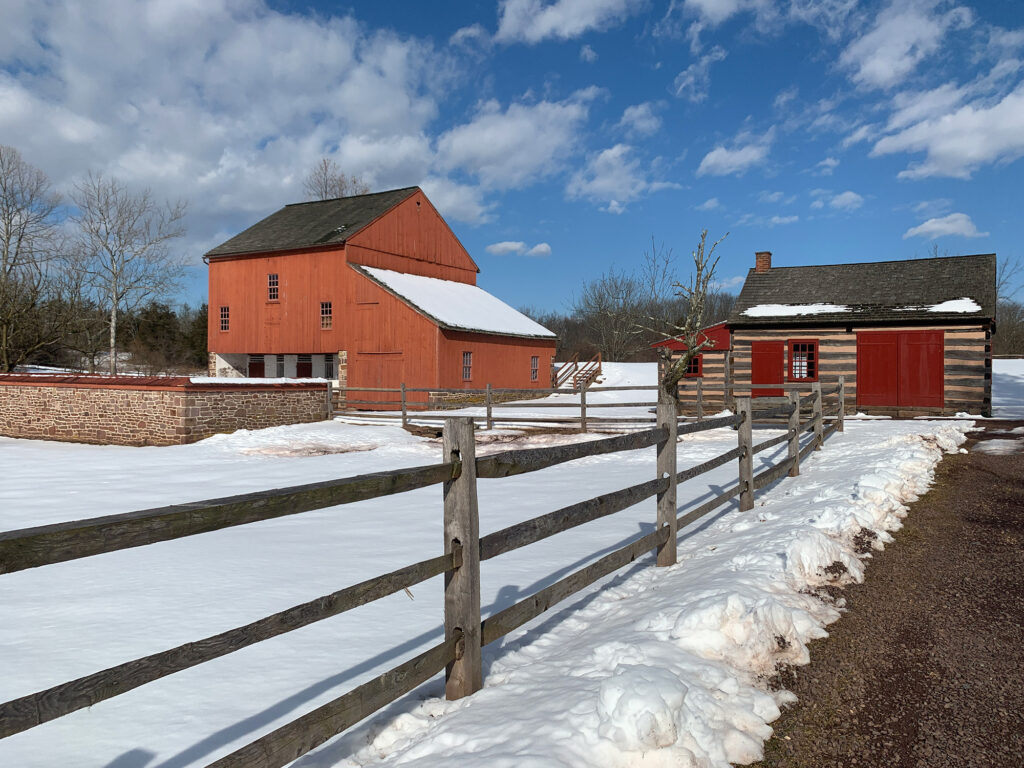 Large reddish orange barn and log cabin against a field of snow at the Daniel Boone Homestead 