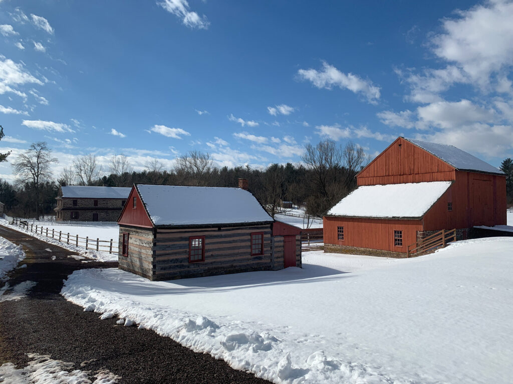 log cabin and a large reddish orange barn in a snowy field at Daniel Boone Homestead