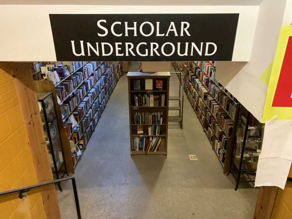 Sign reading "Scholar Underground" above a staircase leading to a basement filled with bookshelves