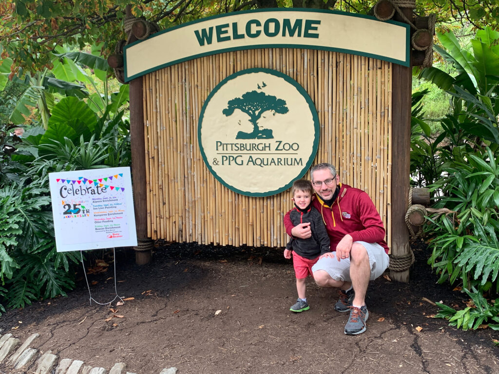 Man with small boy standing in front of a wooden welcome sign at the Pittsburgh Zoo