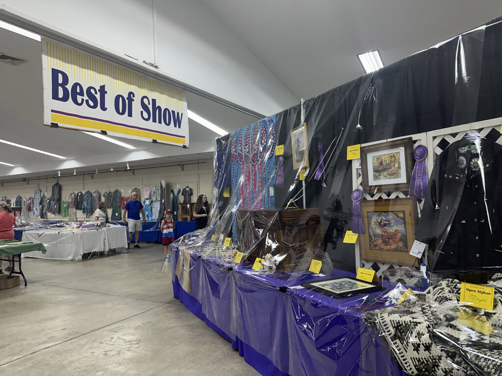 large sign reading "Best of Show" above a table with purple tablecloth and various baked goods and handmade items