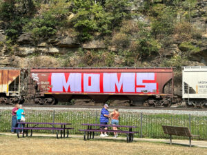 brown hopper car with the words "MOMS" in large block graffiti letters in light purple with red accents