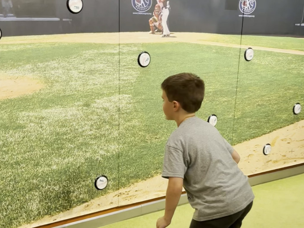 young boy looking at a wall made to look like a Little League field with buttons to push to indicate a hit ball