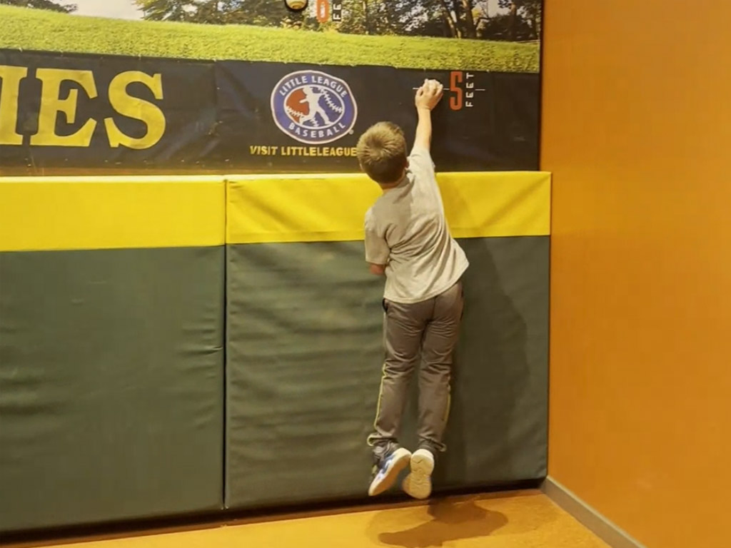 child jumping against a replica outfield wall to touch a baseball at the five-foot mark