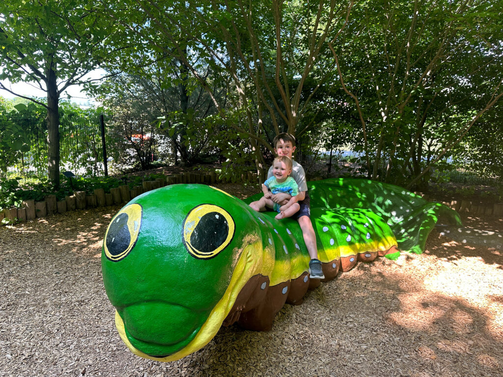 a young boy and a toddler sit on a green caterpillar bench at Penn State Arboretum