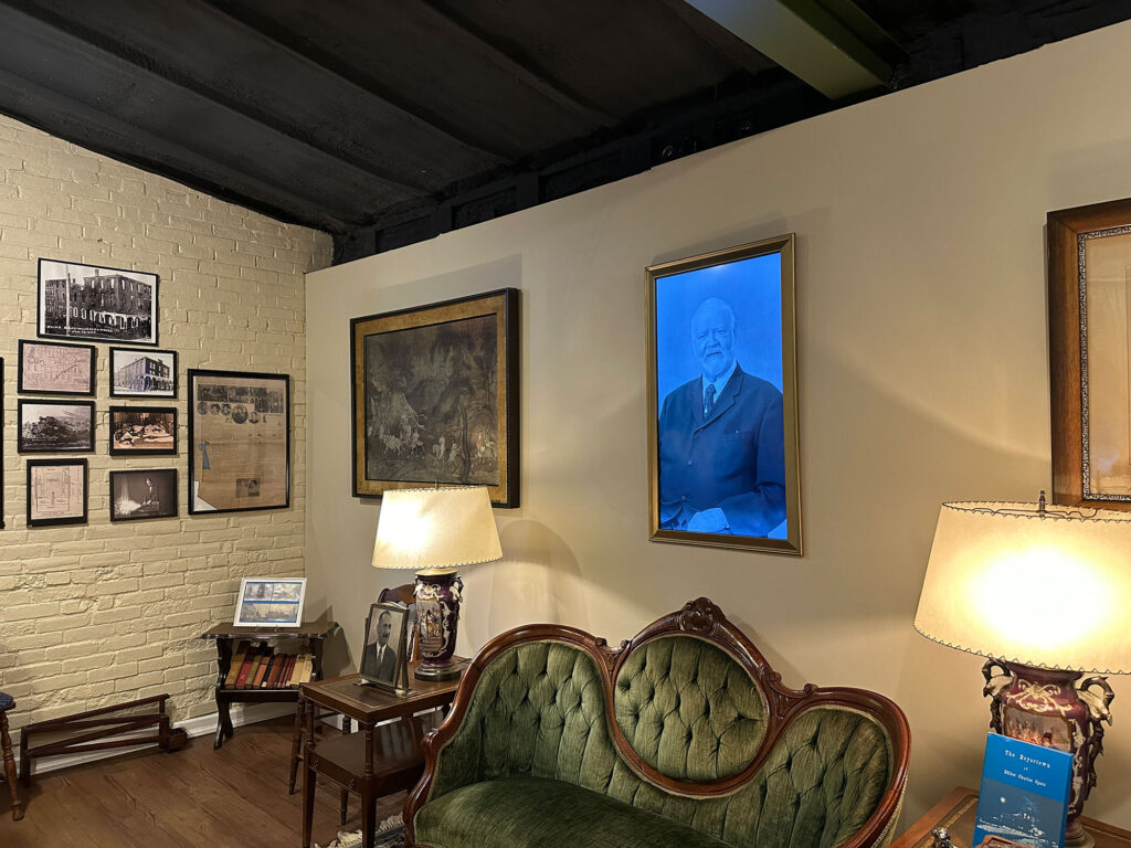 digital portrait on a wall shows a man in early 1900s suit. the picture hangs above a green velvet couch