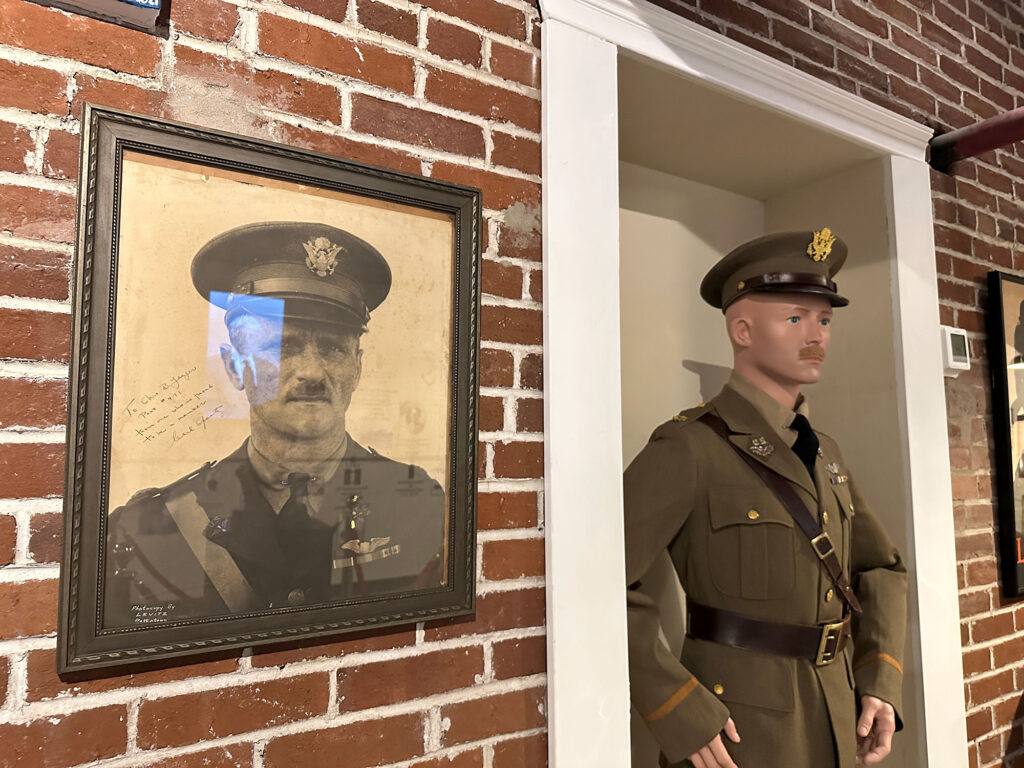 life-size manequin in US Army officer's uniform next to a portrait on the wall of the person it is depicting