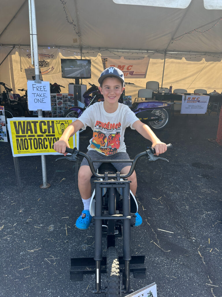 young boy on a stationary motorcycle in front of a side that reads "watch for motorcycles"
