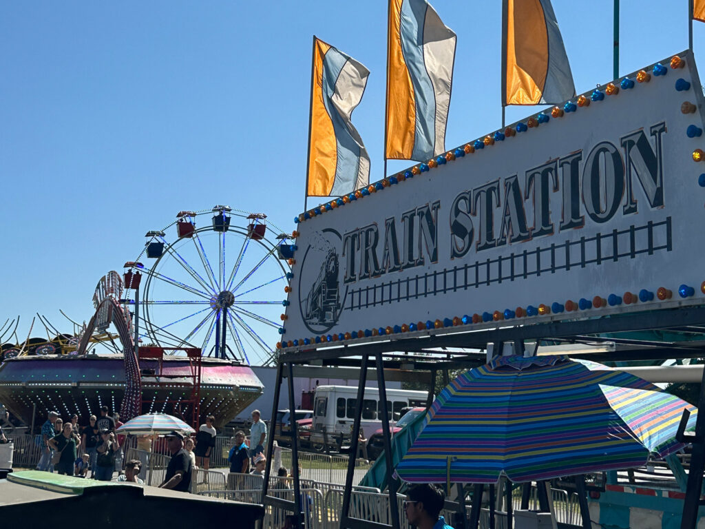 sign reading "train station" above a carnival ride with a Ferris Wheel and flags in the background