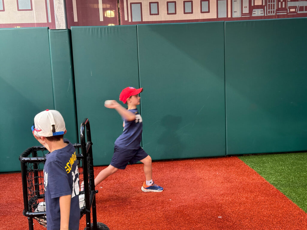 young boy pitching in a replica field with padded green wall