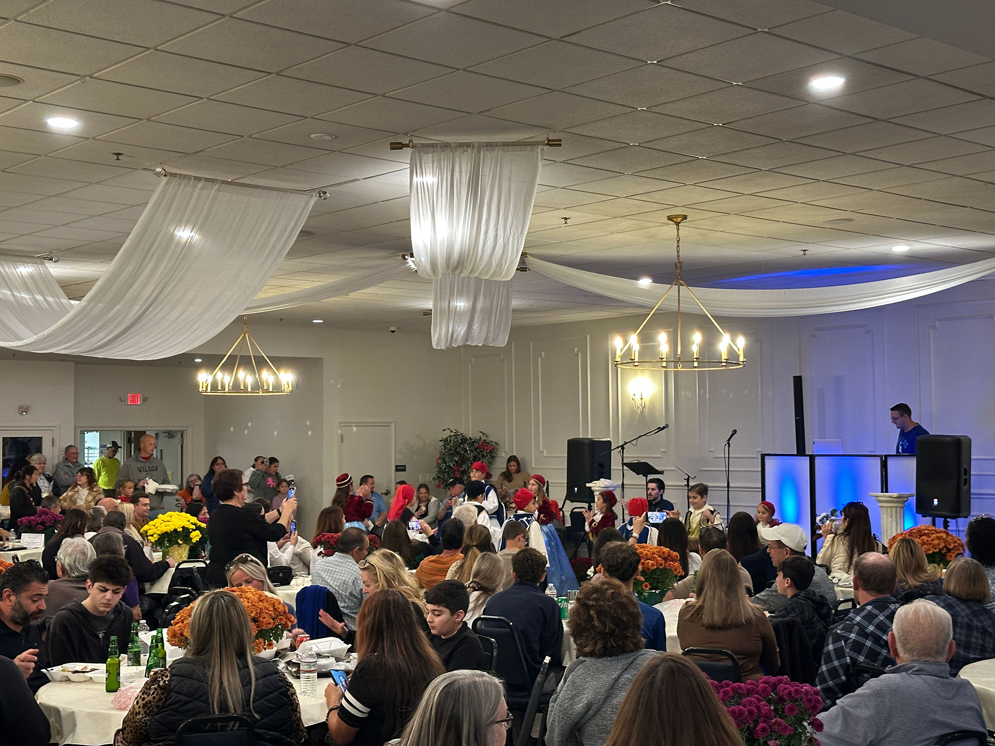 Room full of people sitting at round tables watching children dance in traditional Greek garb.