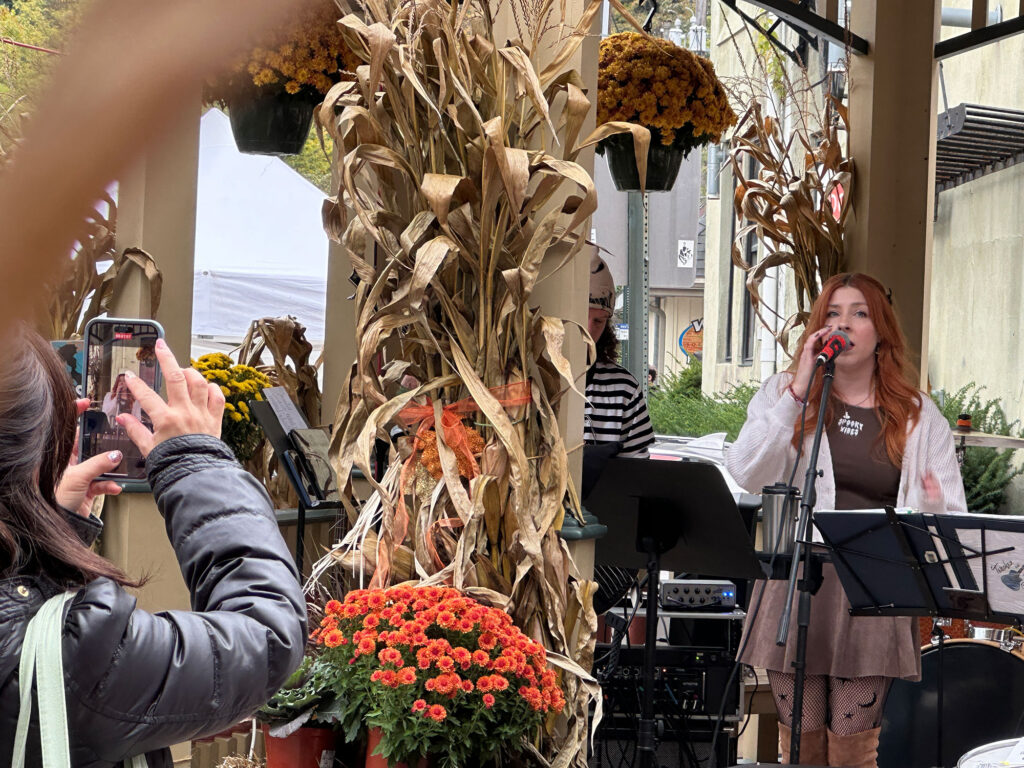 woman singing in a gazebo decorated with corn stalks as another woman takes a photo