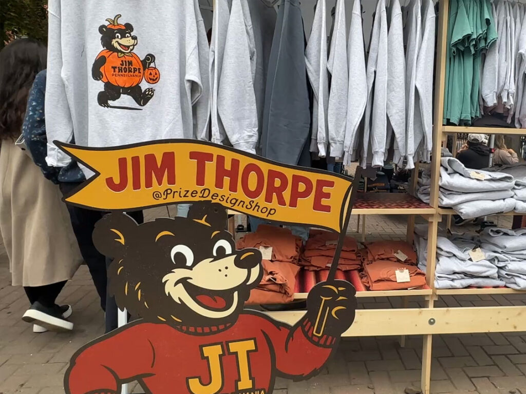 A wood cut-out of a cartoon bear wearing a red sweater with JT in yellow letters holding a banner that says "JIm Thorpe Pennsylvania" stands in front of a t-shirt stand.