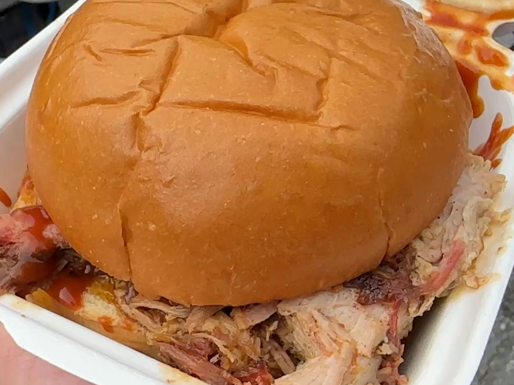An overstuffed pulled pork sandwich sits inside a white clamshell package.