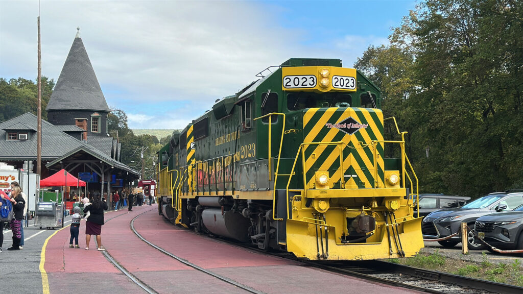 A green diesel locomotive with yellow stripes on the front and the number 2023 above the cab on the side sits next to the Jim Thorpe train station with its witch's hat architecture.