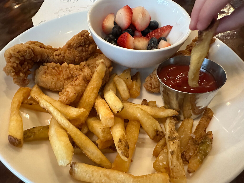 white plate with two chicken tenders, a pile of fries, a cup of fruit and a small cup of ketchup where a hand is dipping a fry.