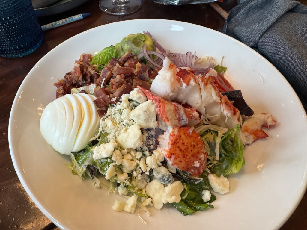 large white plate with a salad topped with half a lobster and a sliced hard-boiled egg