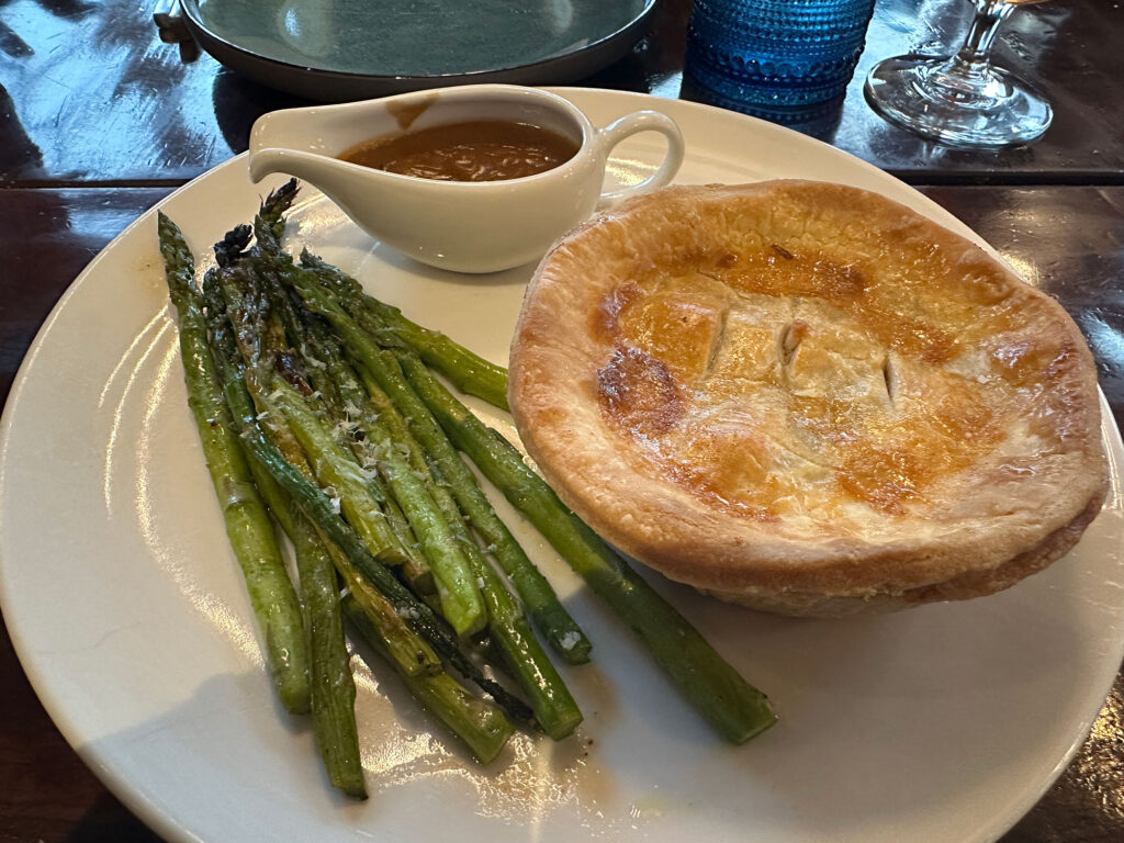 white round plate with an individual sized pie, spears of aspargus, and a small gravy boat with brown gravy