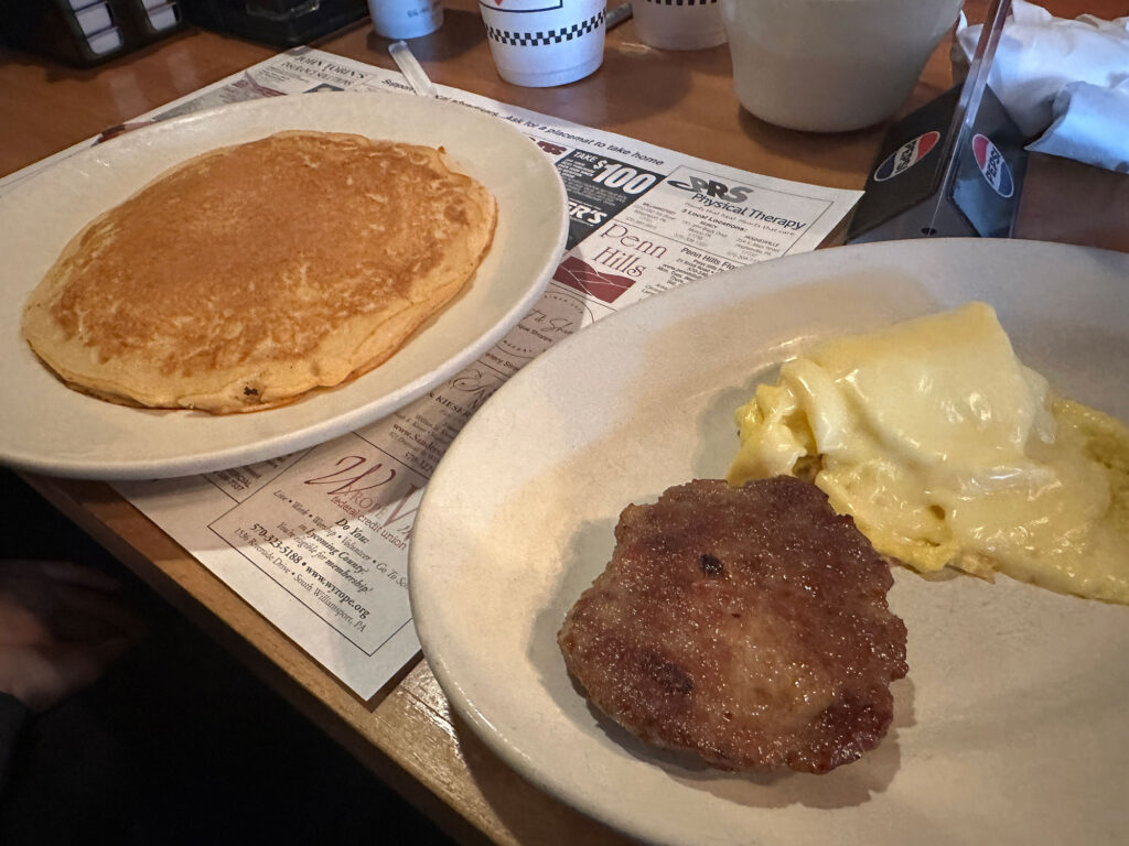 two plates side-by-side, one with a plate-sized pancake, the other with a scrambled egg and a sausage patty