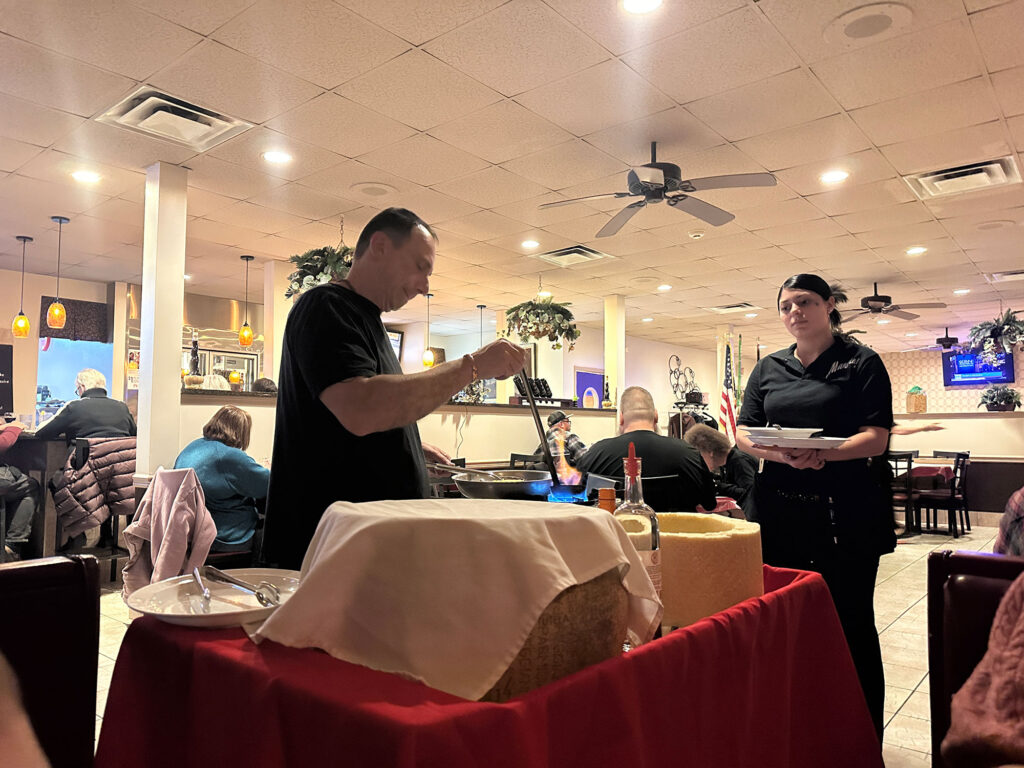 man holding a ladle full of alcohol that has a blue flame coming out of it while a woman stands holding a plate with spaghetti