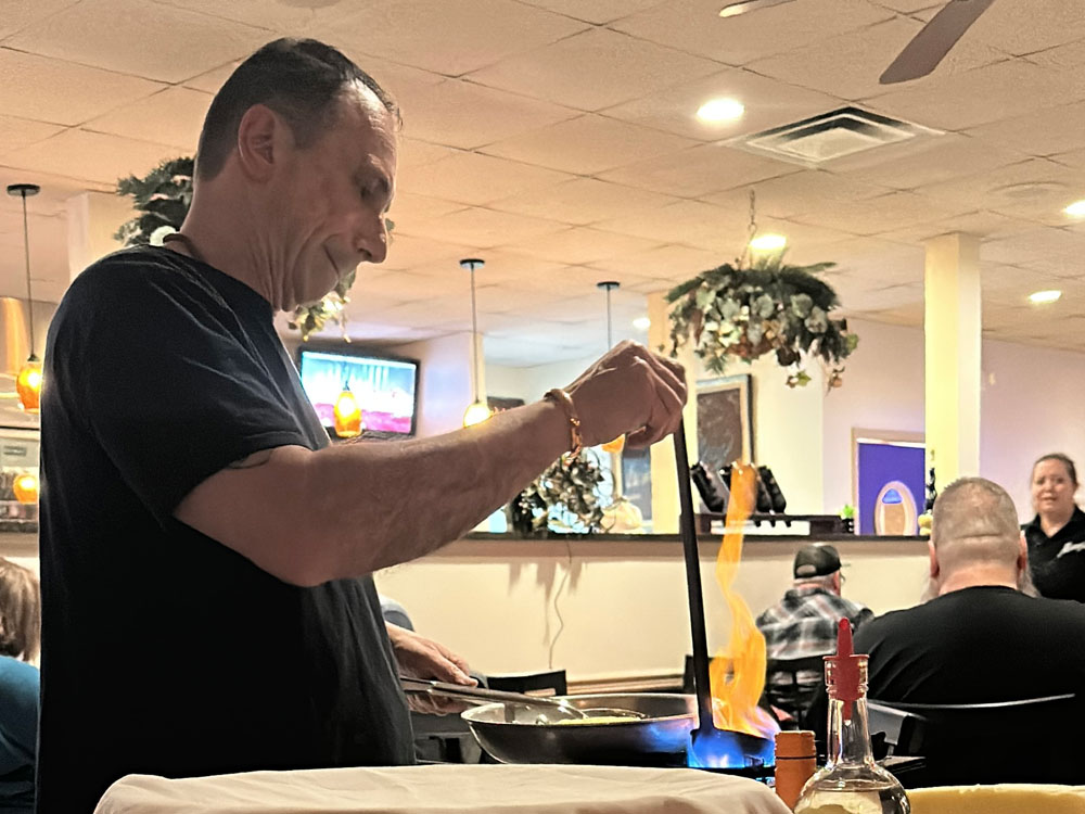 man holding a ladle full of grappa over a fire as orange flames come out of the alcohol