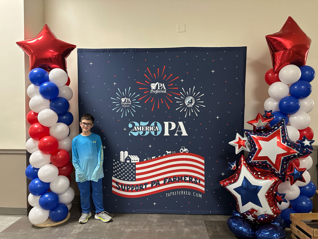 young boy in a light blue long-sleeve shirt and blue pants stands in front of a blue backdrop with an American flag flanked by red, white and blue balloon towers