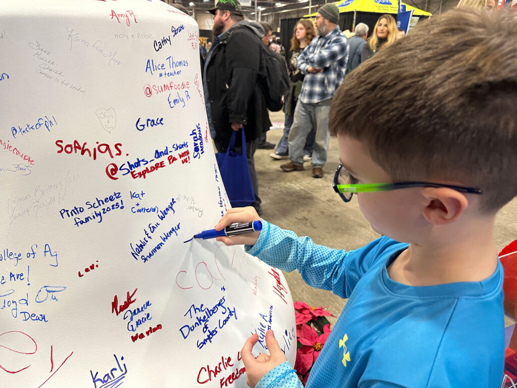 young boy uses a blue marker to sign his name on a white replica of the Liberty Bell