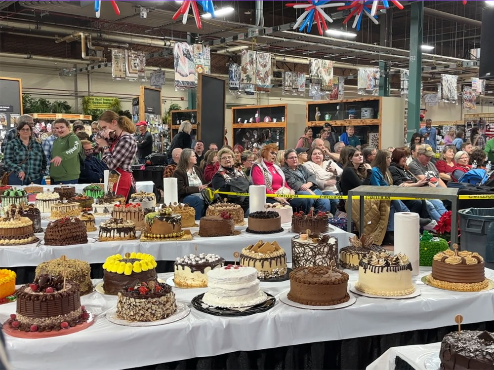 tables with various chocolate cakes in front of a crowd of people sitting watching a stage.
