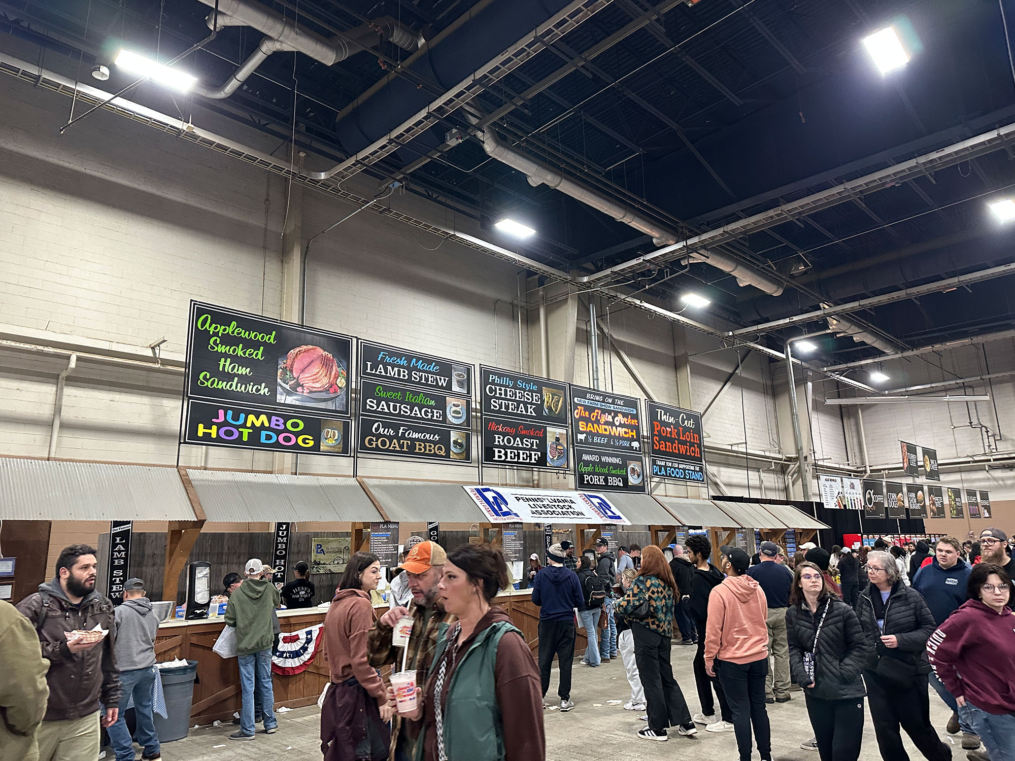 large crowd of people walking and standing in line near long food stand with pictures of various food items over head