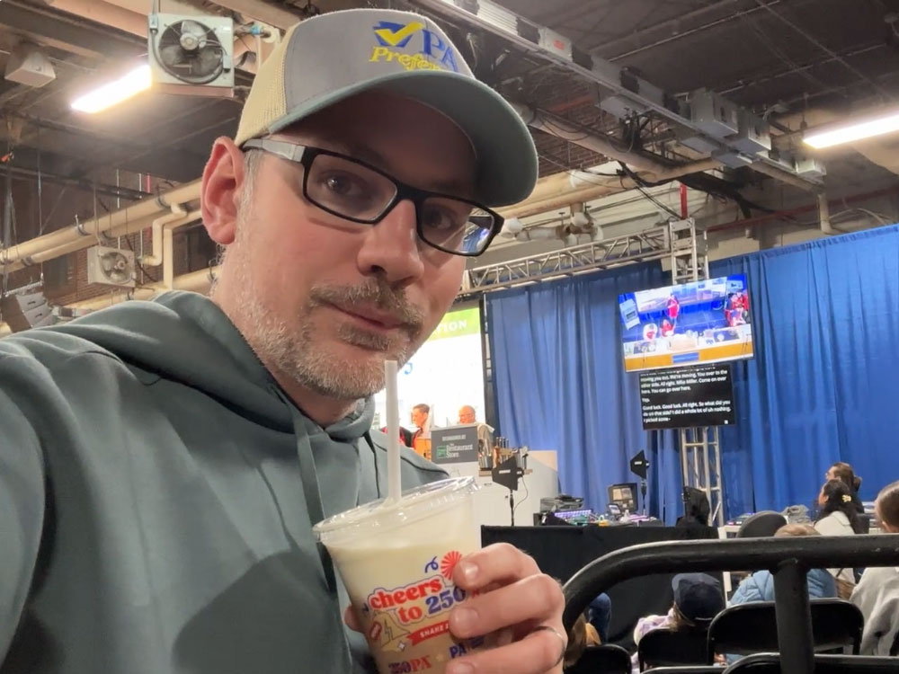 a man wearing a green-gray hoodie with a matching hat bearing the words "PA Preferred" taking a selfie holding a half-chocolate, half-vanilla milkshake.