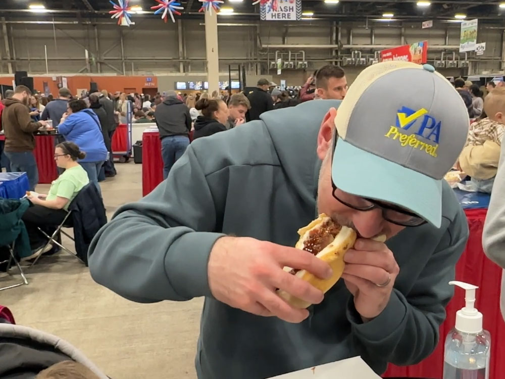 a man wearing a hat with the words "PA Preferred" hunches over to take a bite from a sandwich.