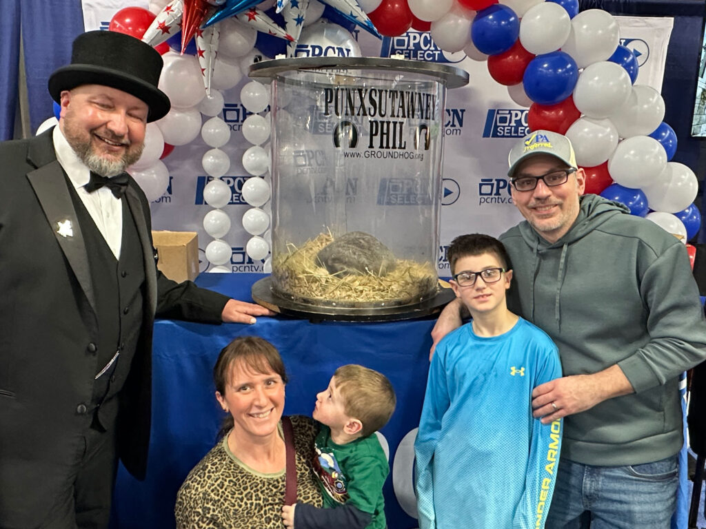 a family with a mom, dad, and two young sons standing with a man in a top hat next to a clear aquarium-like structure with a groundhog inside.