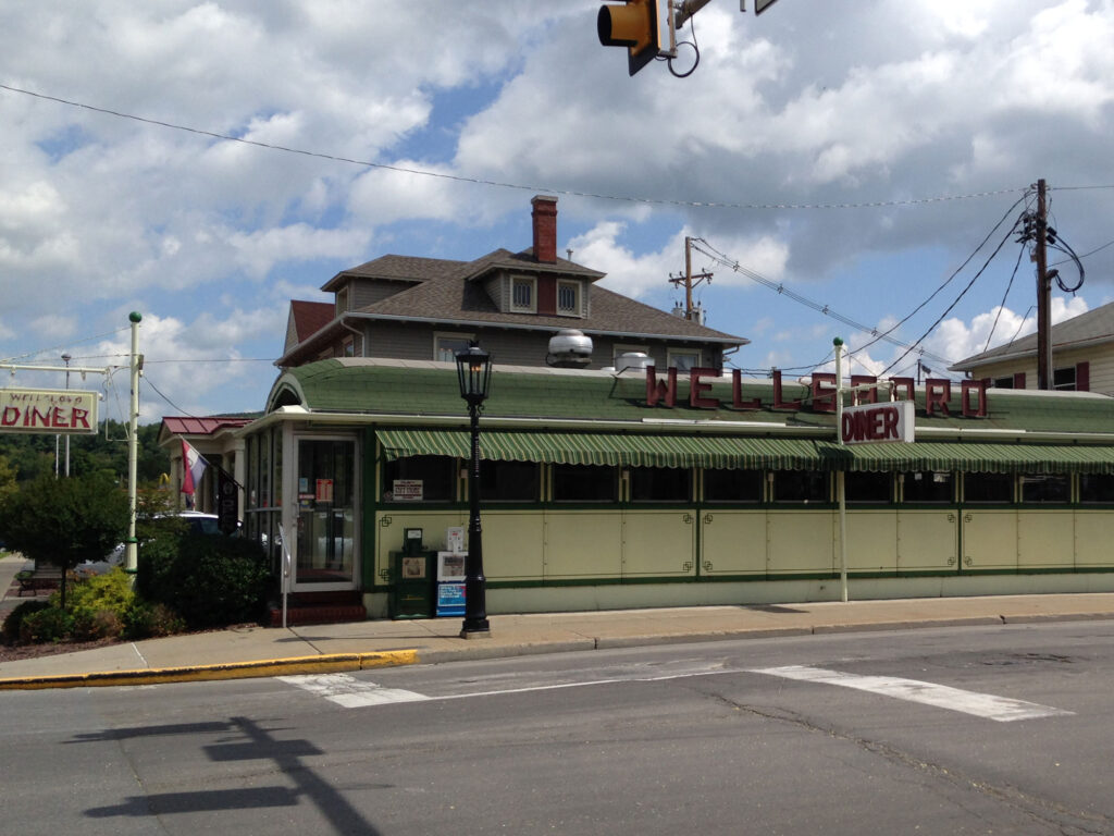 Old-fashioned diner with green roof and yellow-green walls and red letters on the roof reading "WELLSBORO". 