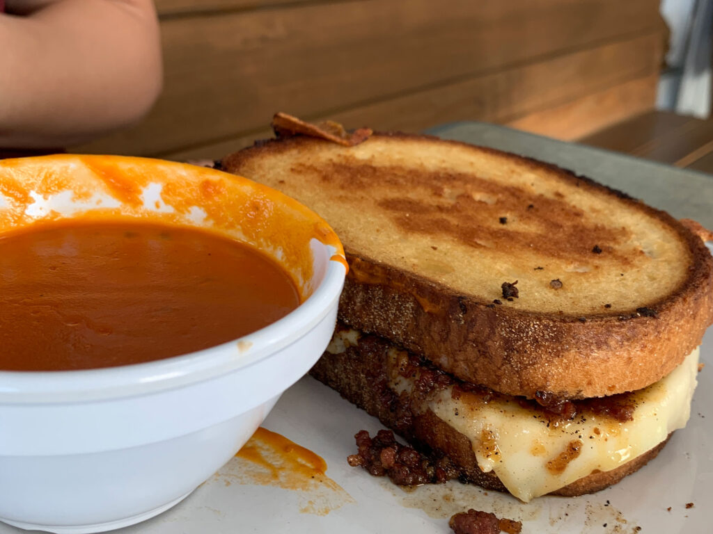 plate with a white bowl of tomoato soup next to a grilled cheese sandwich with bacon jam and cheese oozing out the side