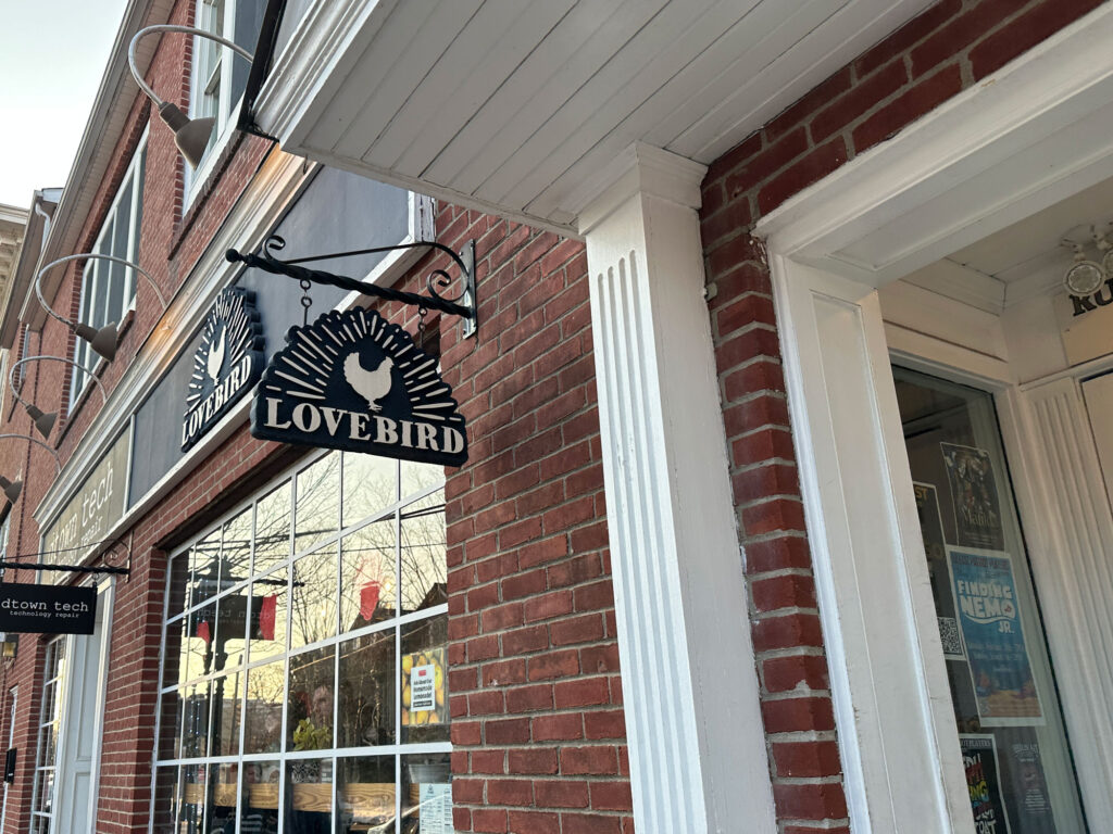 brick building with white trim and a black sign hanging next to the door ith a white chicken and the word "Lovebird"
