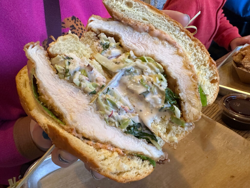 hands holding a sandwich cut in half to show layers of fried chicken, sauce, coleslaw and cheese