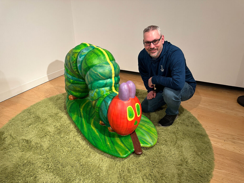 man in a blue hoodie leaning down next to a sculpture of the Very Hungry Caterpillar
