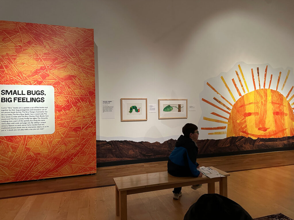 young child sitting on a bench in front of a wall that has a sign reading "Small bugs, big feelings" and a large sun in the style of Eric Carle