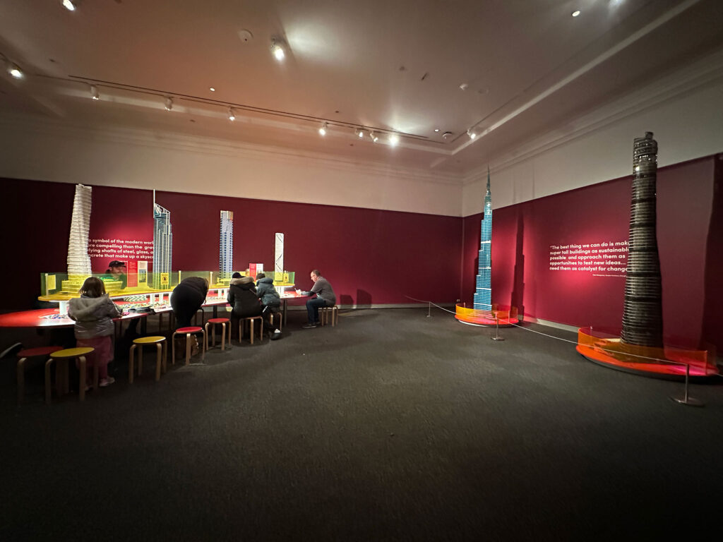 large galllery space at the Reading Public Museum with red walls - on the left, a kidney bean-shaped table where people are building with LEGOS and on the right are two models of skyscrapers built out of LEGOS